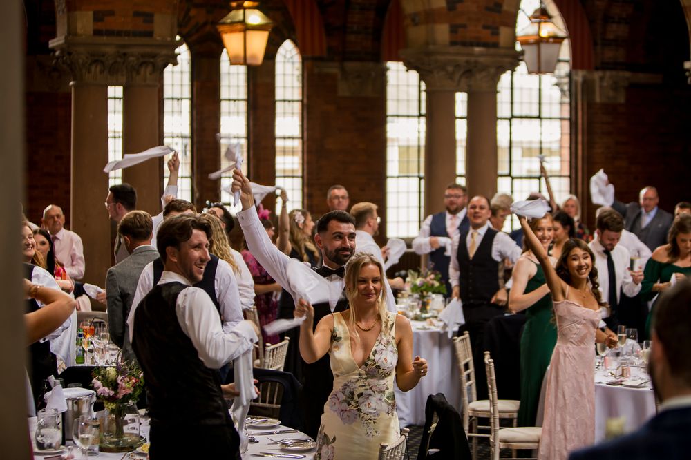 Wedding guests wave napkins in celebration inside historic venue with tall windows.