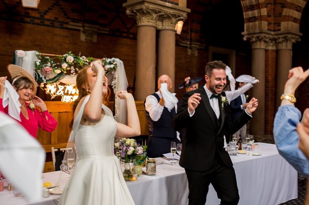 Wedding celebration with guests dancing and cheering in an elegant venue with ornate columns and arches.