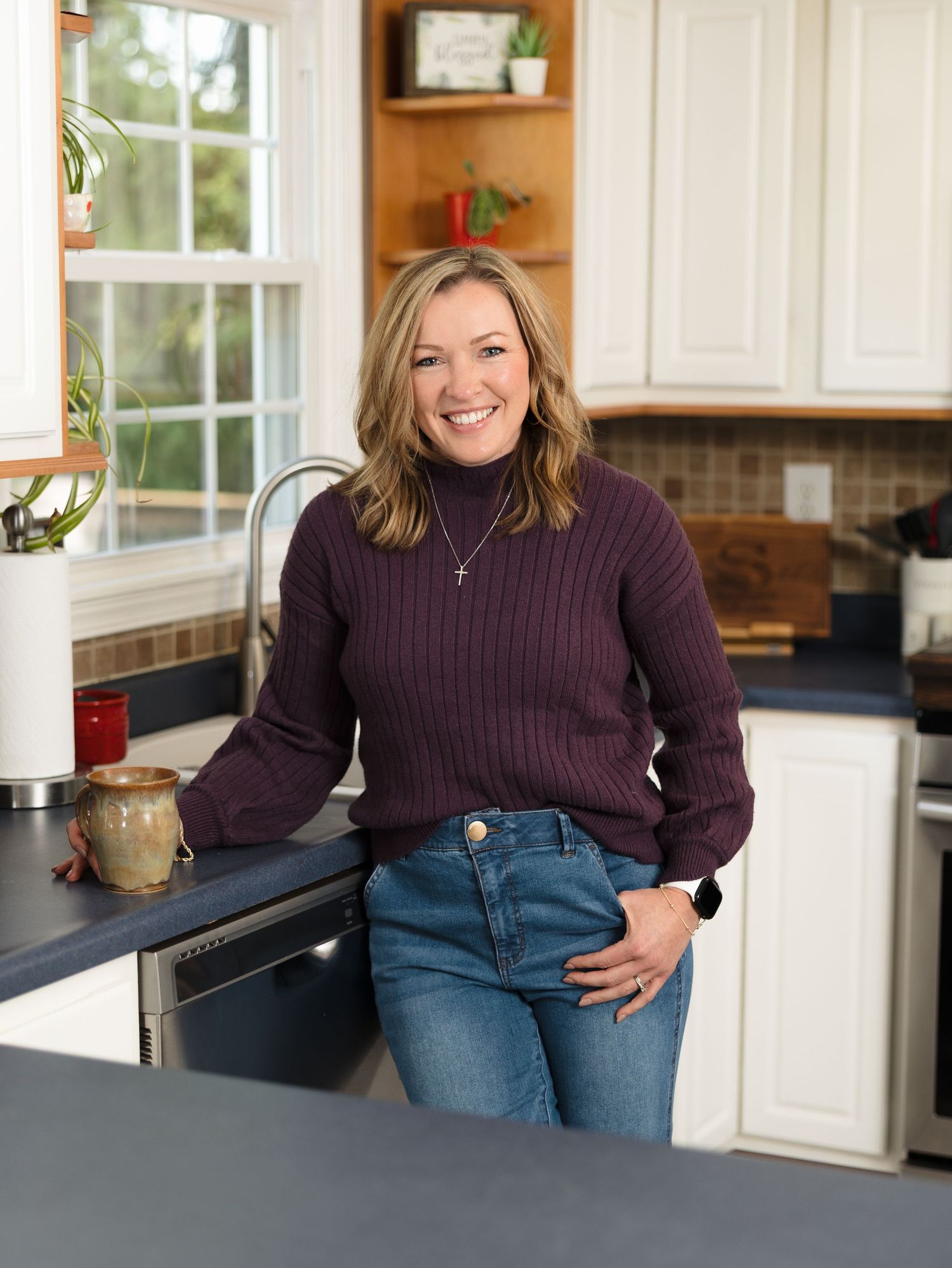 Person in purple sweater and jeans sitting casually on kitchen counter in bright modern white kitchen.