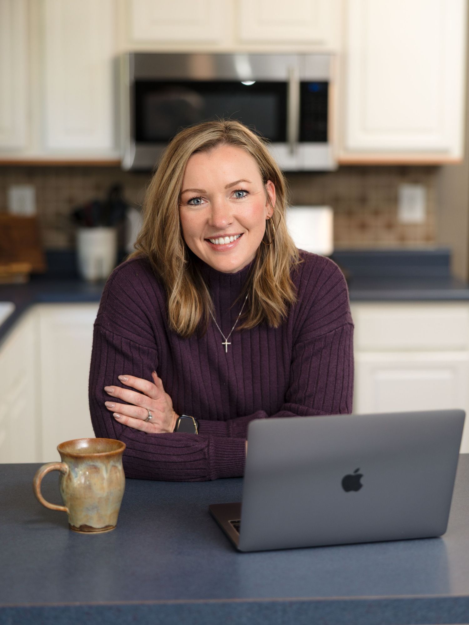 Someone in a purple sweater works from home at a kitchen counter with a MacBook laptop and ceramic mug nearby.