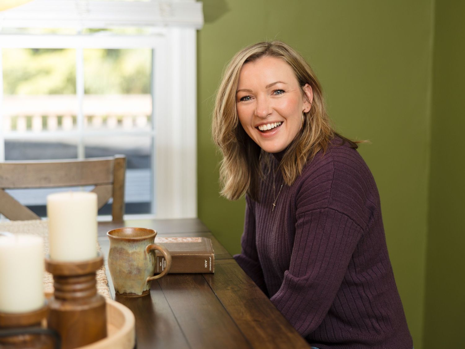 A person in a brown sweater stands in a modern kitchen with green walls and white cabinets.
