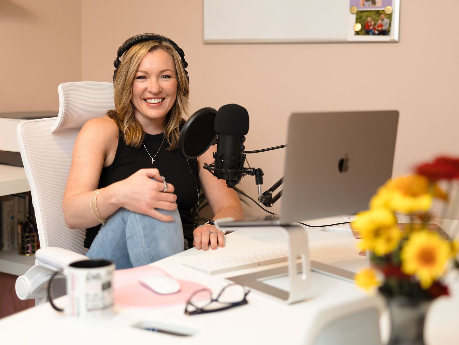 A person in a white office setting smiles while working at a desk with a laptop and yellow flowers.