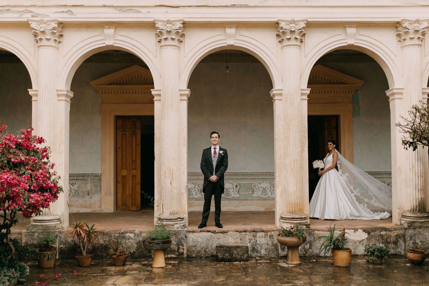 Newlyweds stand on outdoor stairs with black metal railings and pink flowers in bloom at the edges.