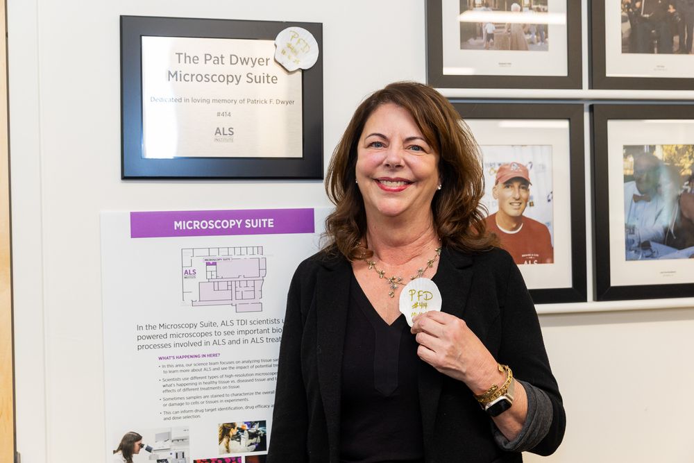 Two people stand smiling in a gallery or office space with framed photos and informational displays on the wall.