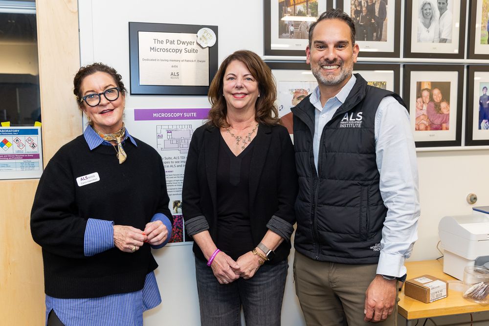 Three healthcare professionals stand together smiling in an office with framed photos on the wall behind them.