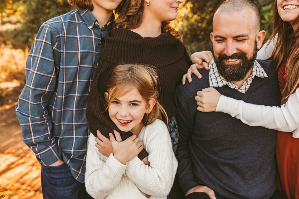 A joyful family laughs together during an outdoor autumn photoshoot with golden sunlight filtering through trees.