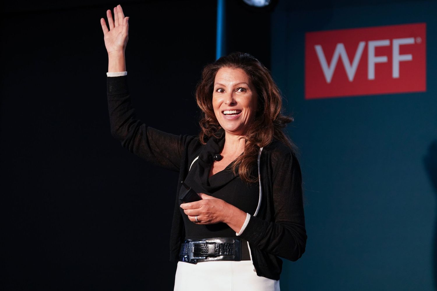 A presenter in a black jacket waves to the audience while standing at a podium with WFF logo visible in the background.