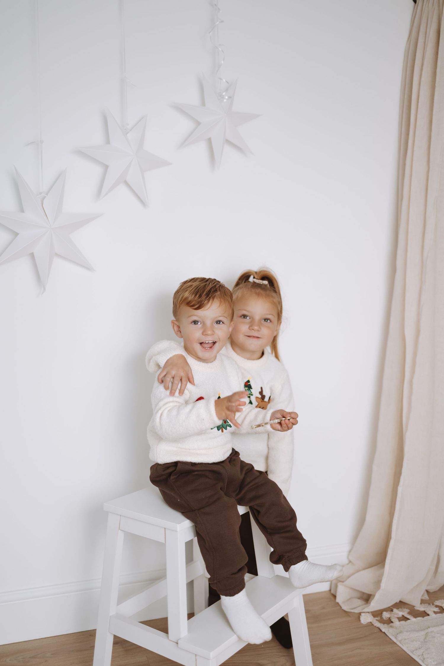 Two children in white sweaters sit together on a white stool against a wall decorated with star ornaments.