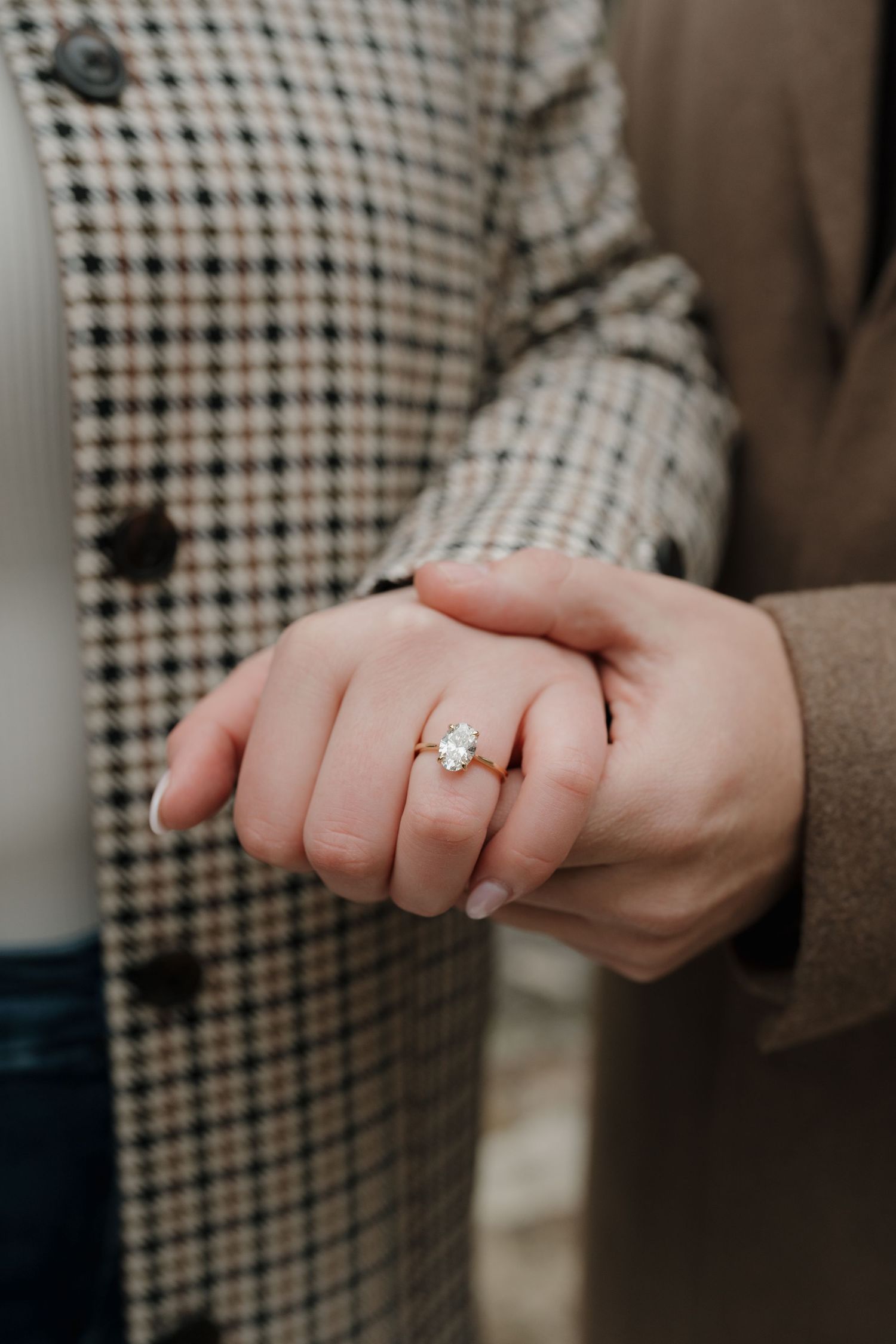 Close-up view of hands displaying an elegant oval-cut diamond engagement ring against a plaid jacket background.