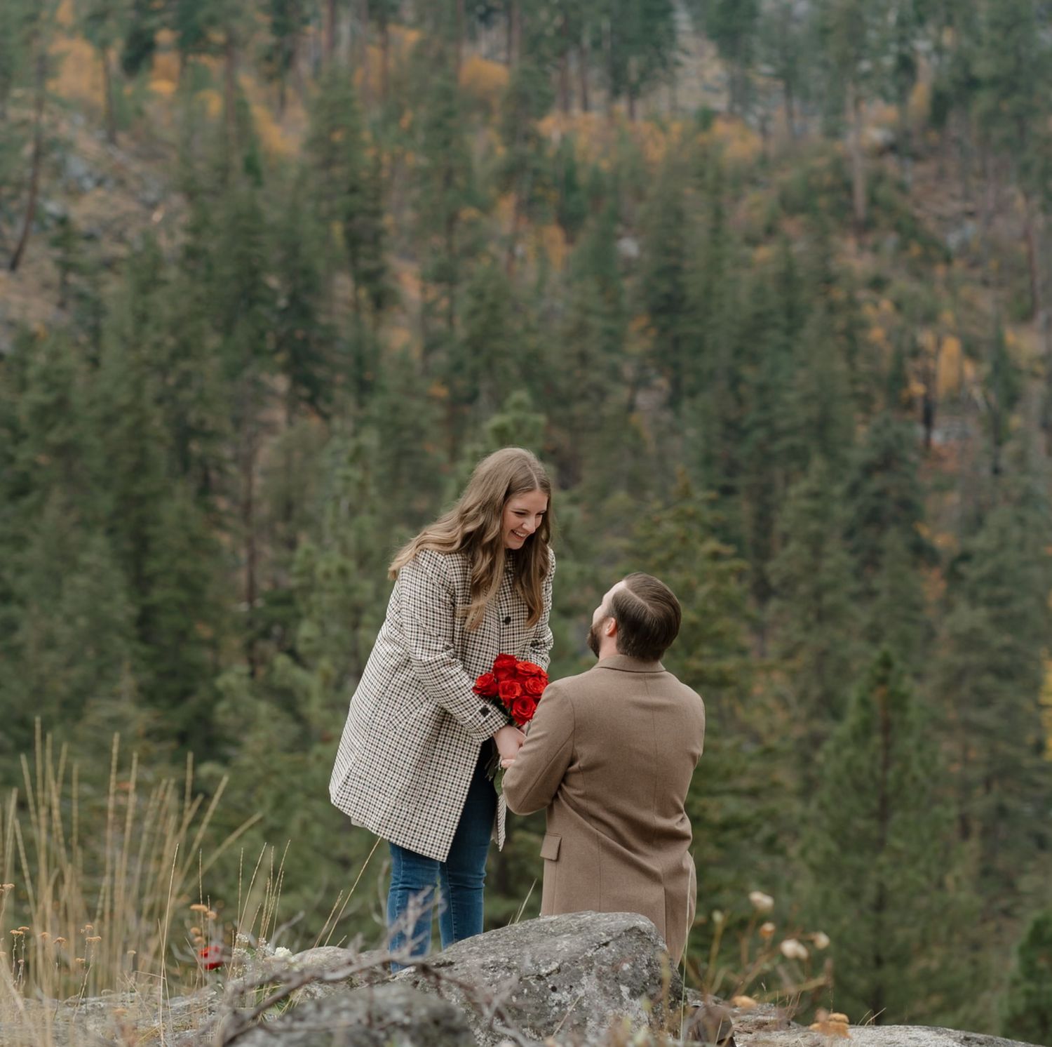 Two people share an intimate moment on a rock overlook surrounded by evergreen forest in autumn.