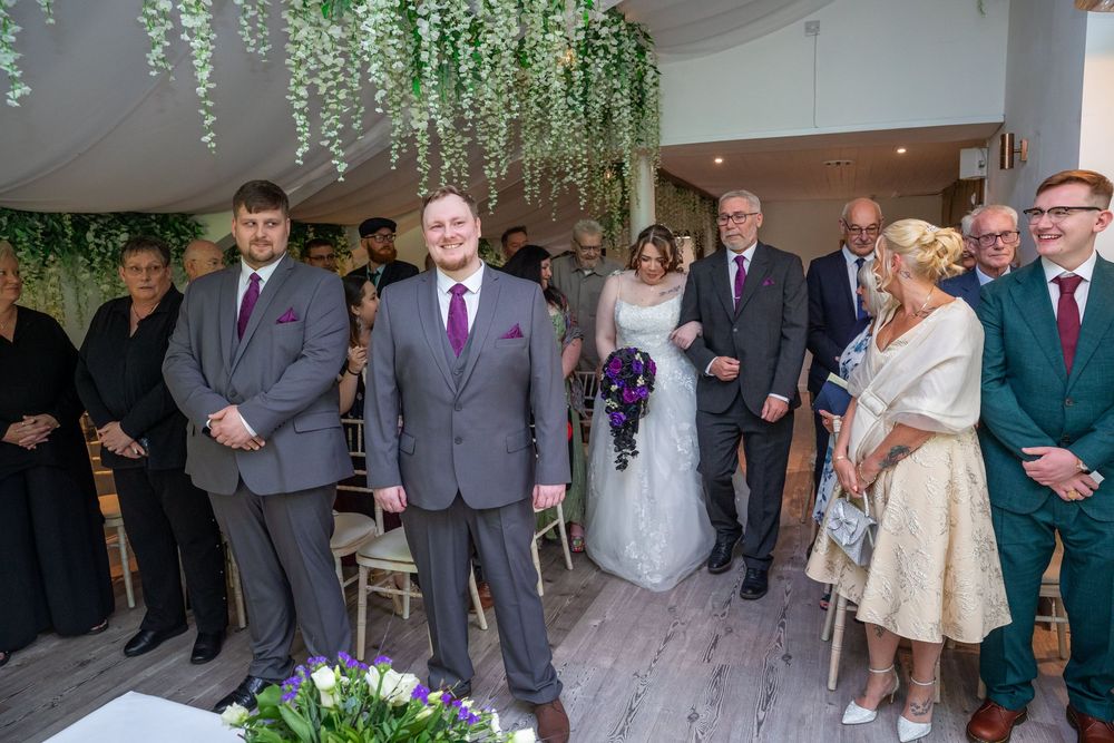 Groom smiles as bride walks down the aisle in a bright modern wedding ceremony venue with floral decorations.