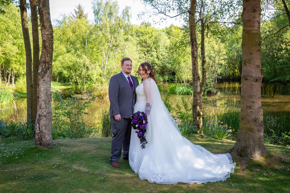 Bride and groom pose together in a lush green woodland setting beside a tranquil lake on their wedding day.