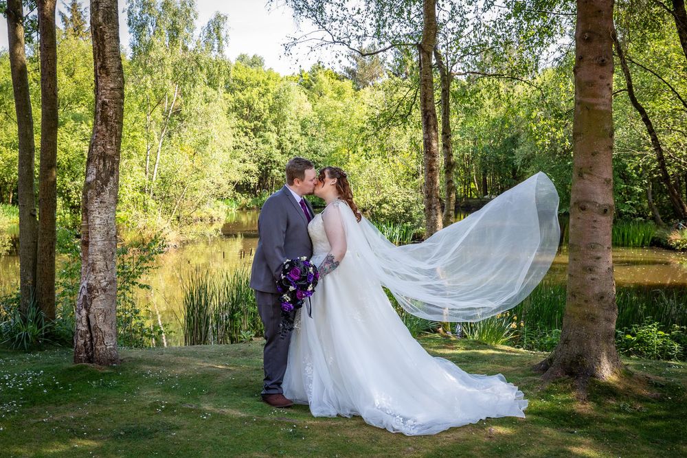 Bride and groom share a kiss as the bride's veil flows beautifully in the breeze by a sunlit woodland lake.