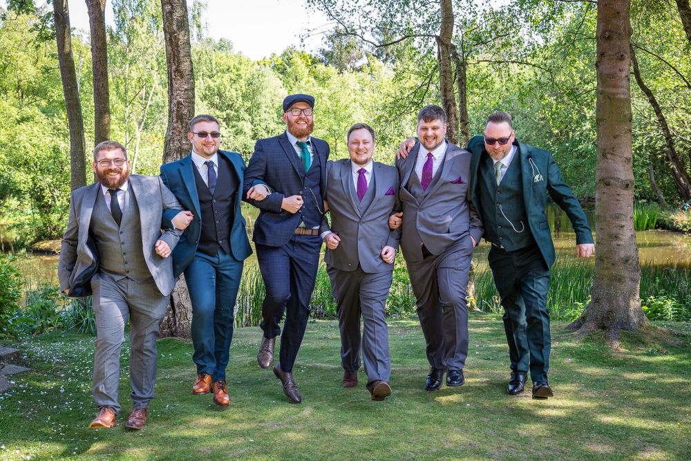 Six groomsmen in coordinating suits laugh and run together through a sunny woodland setting near a lake.