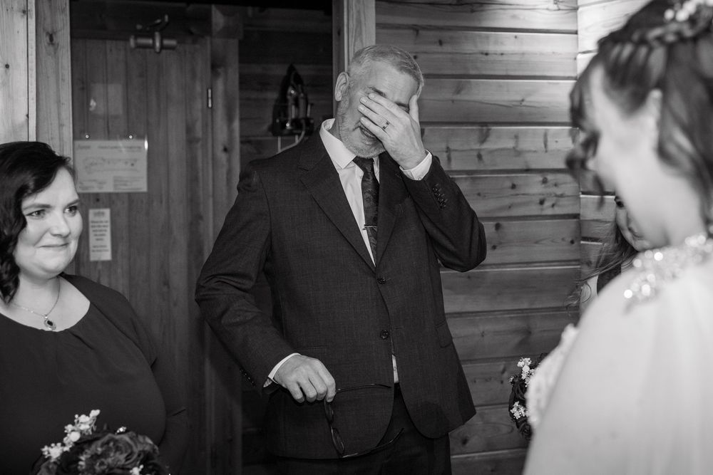 Emotional father wipes tears seeing his daughter in her wedding dress in a black and white candid photo.