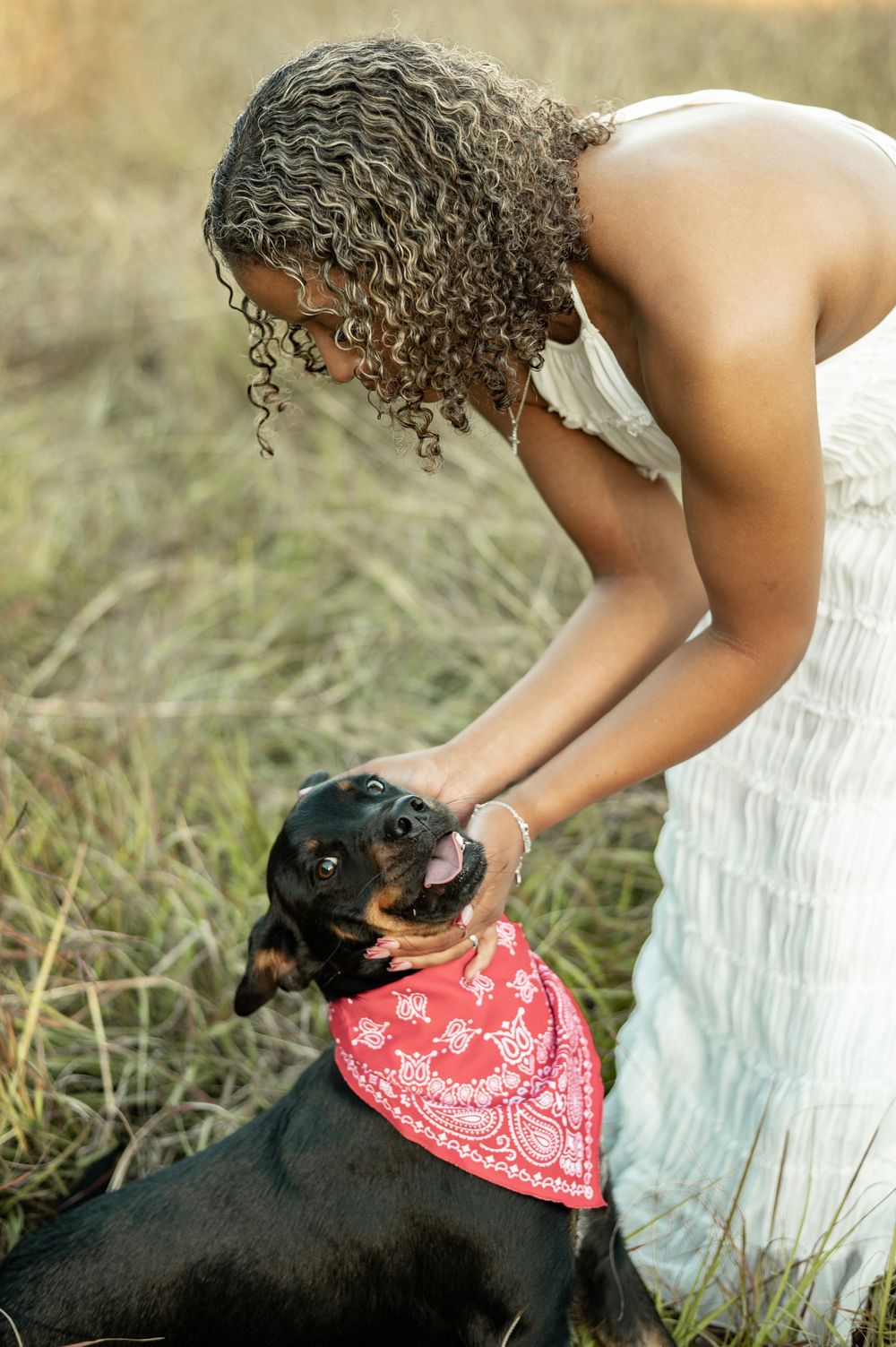 A person in a white dress pets a black dog wearing a red bandana in a grassy outdoor setting.