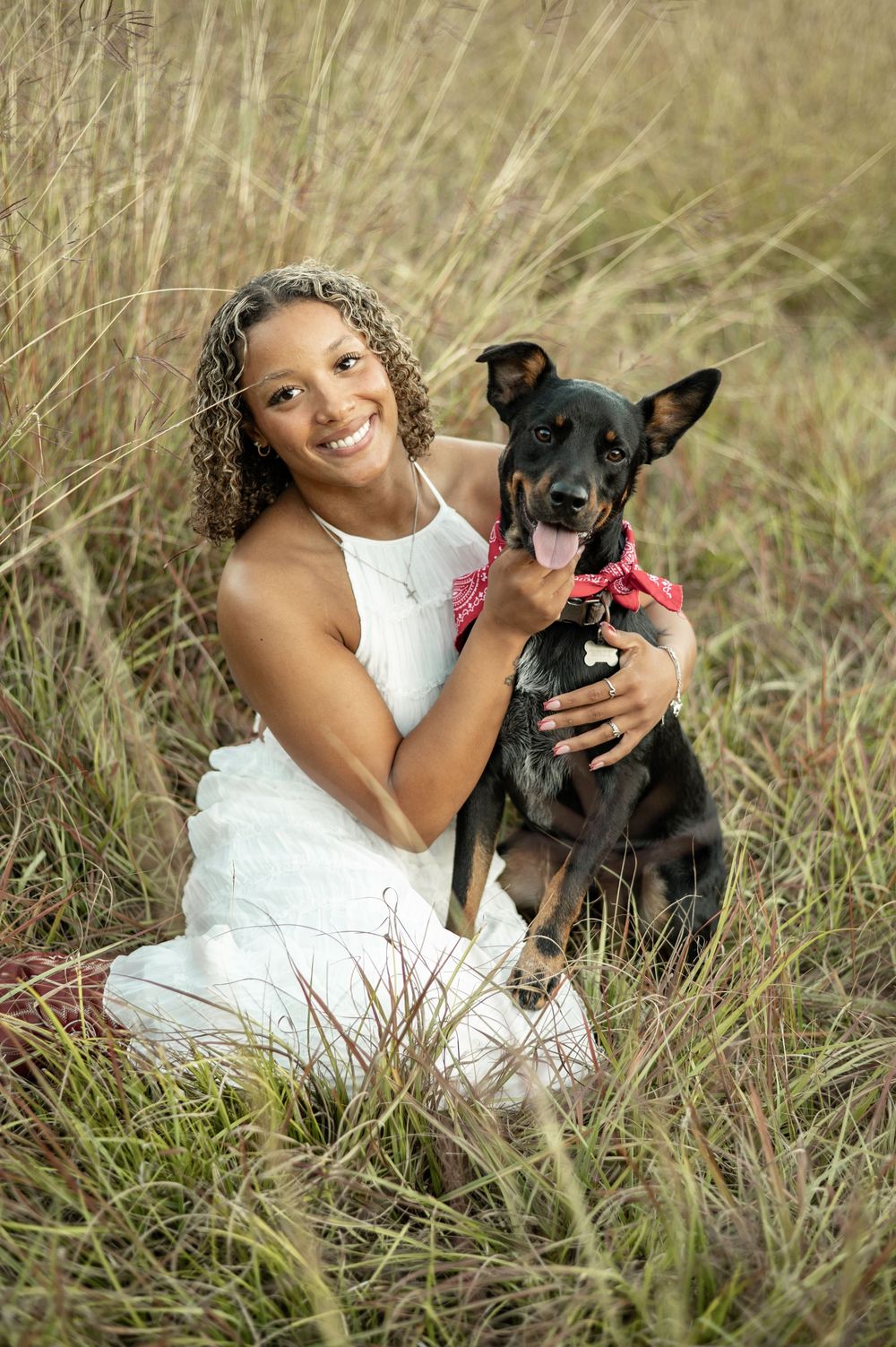 A person in a white dress sits in tall grass with a black dog during an outdoor photo session.