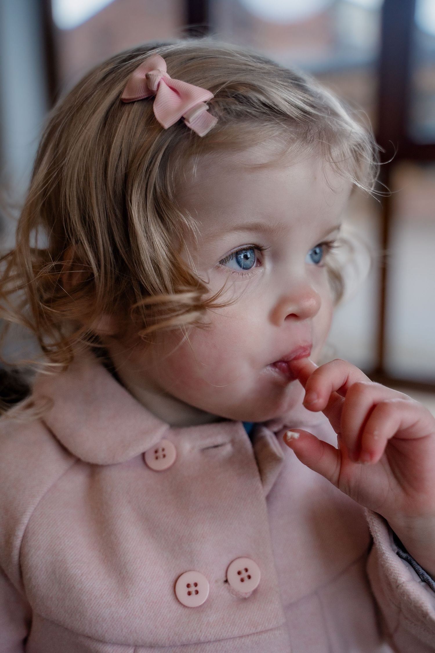 Young child wearing a pink coat with decorative buttons gazes thoughtfully while wearing a bow hair accessory.