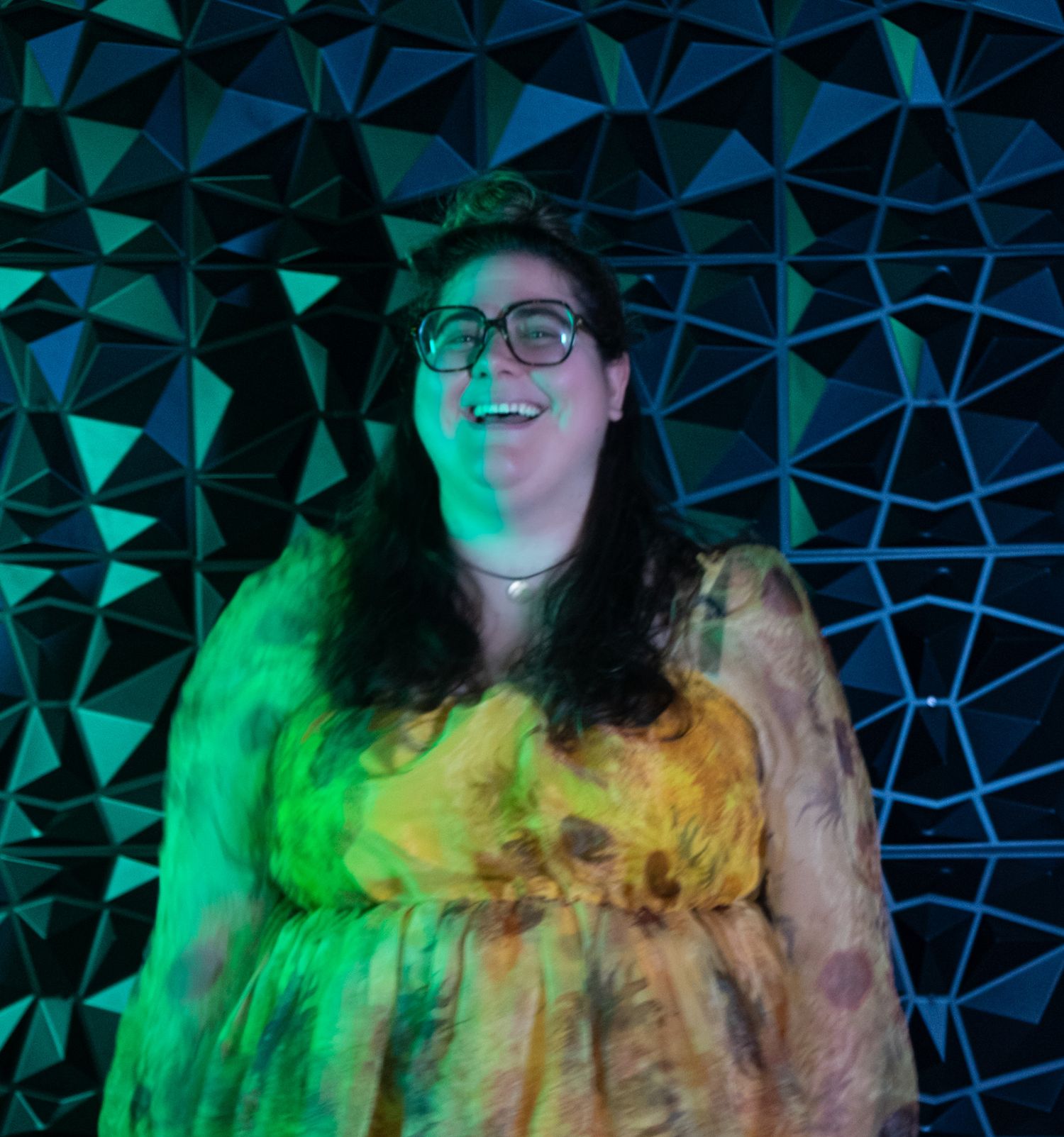 Person with long hair illuminated by vibrant green and blue lighting against a geometric patterned background.