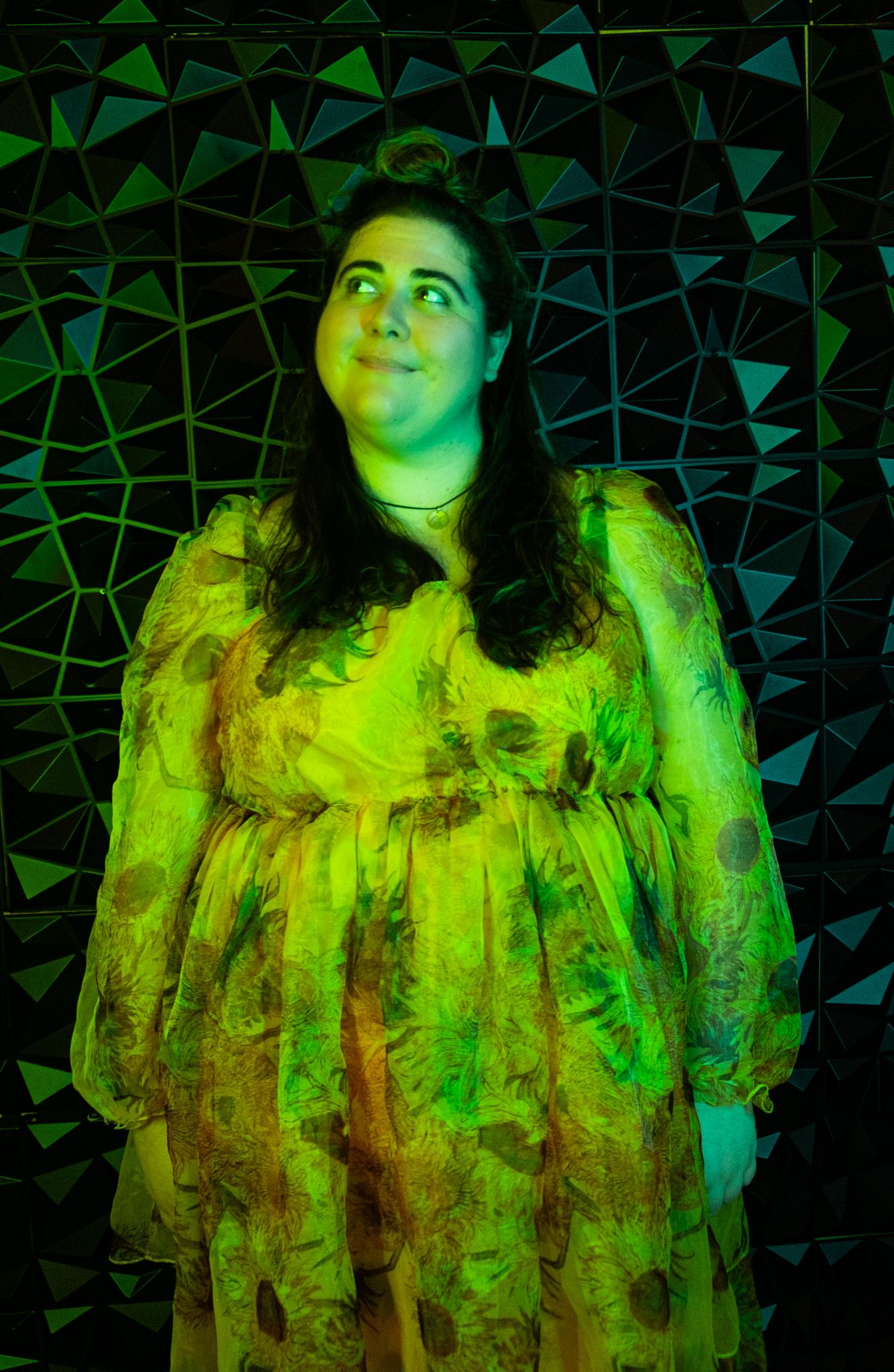 Figure in a flowy dress illuminated by dramatic green lighting against a geometric patterned background.