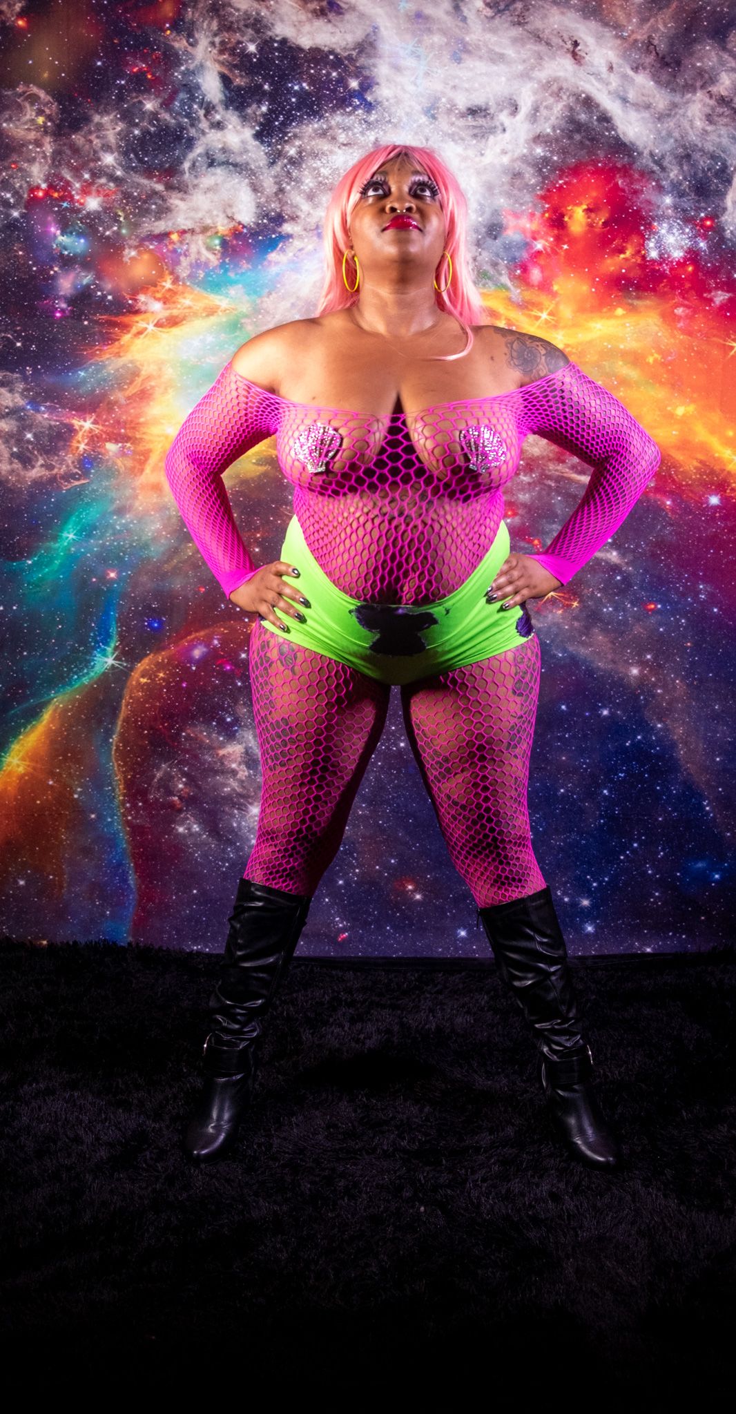 A figure in pink fishnet and neon green top poses dramatically against a cosmic galaxy background with vibrant colors.