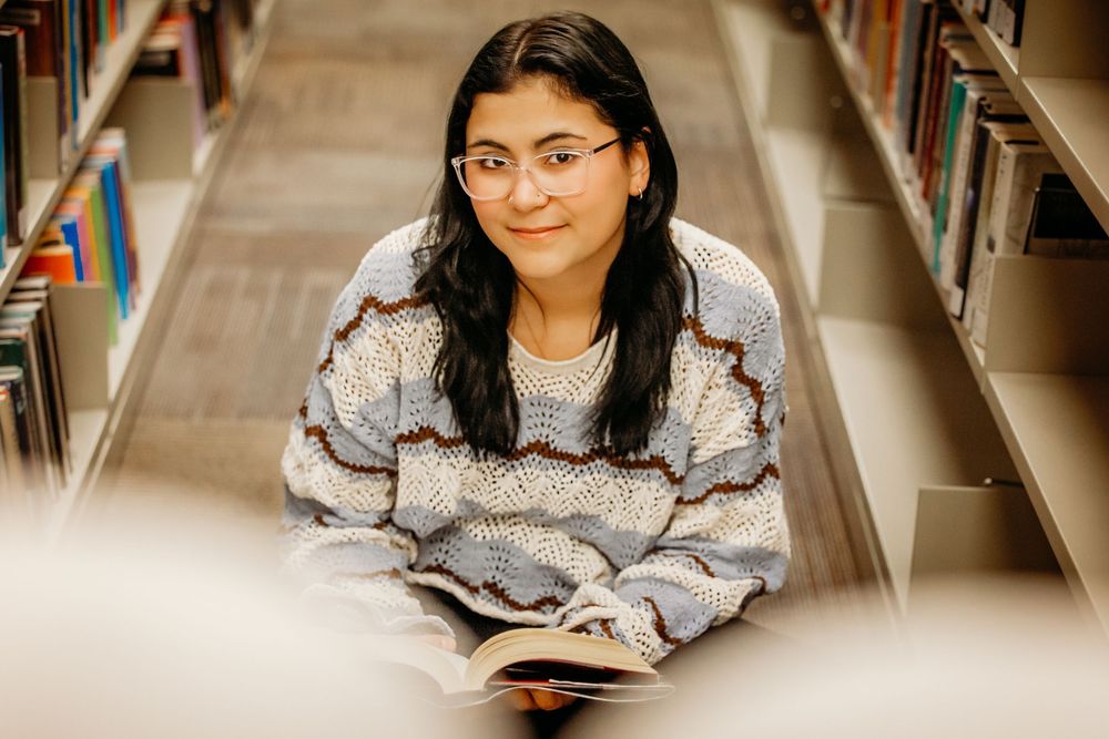 Student in glasses studies at library desk surrounded by books and bookshelves.