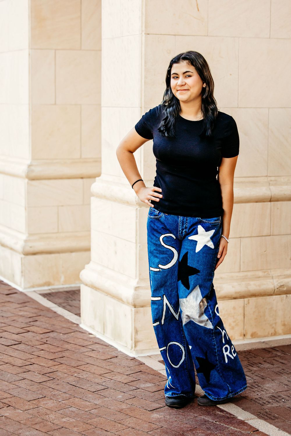 Woman in black t-shirt and custom painted blue jeans with moon and star designs poses against white pillars.