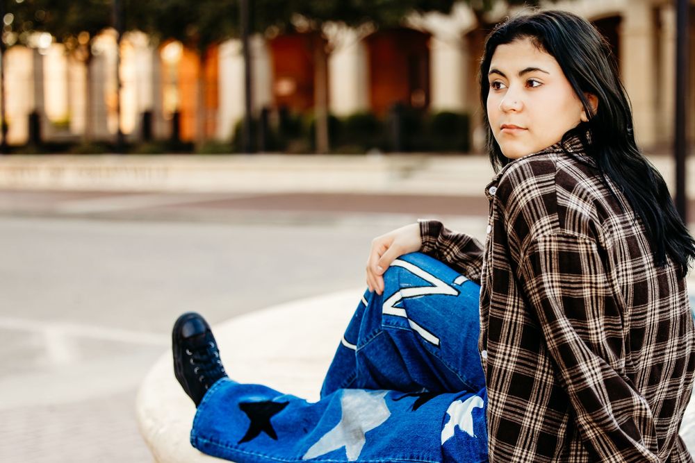 A person wearing plaid and denim sits on a bench with blue bags and shopping items on a sunny day.