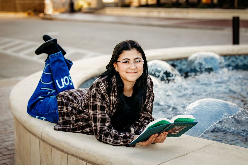 A person in a plaid shirt lies on a white surface while reading a book in a relaxed pose.