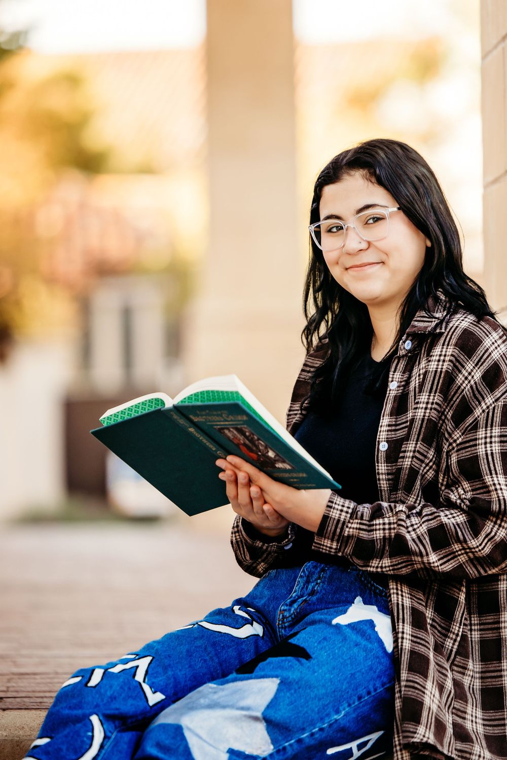 A person wearing a plaid shirt and jeans sits outdoors reading a green book while smiling.