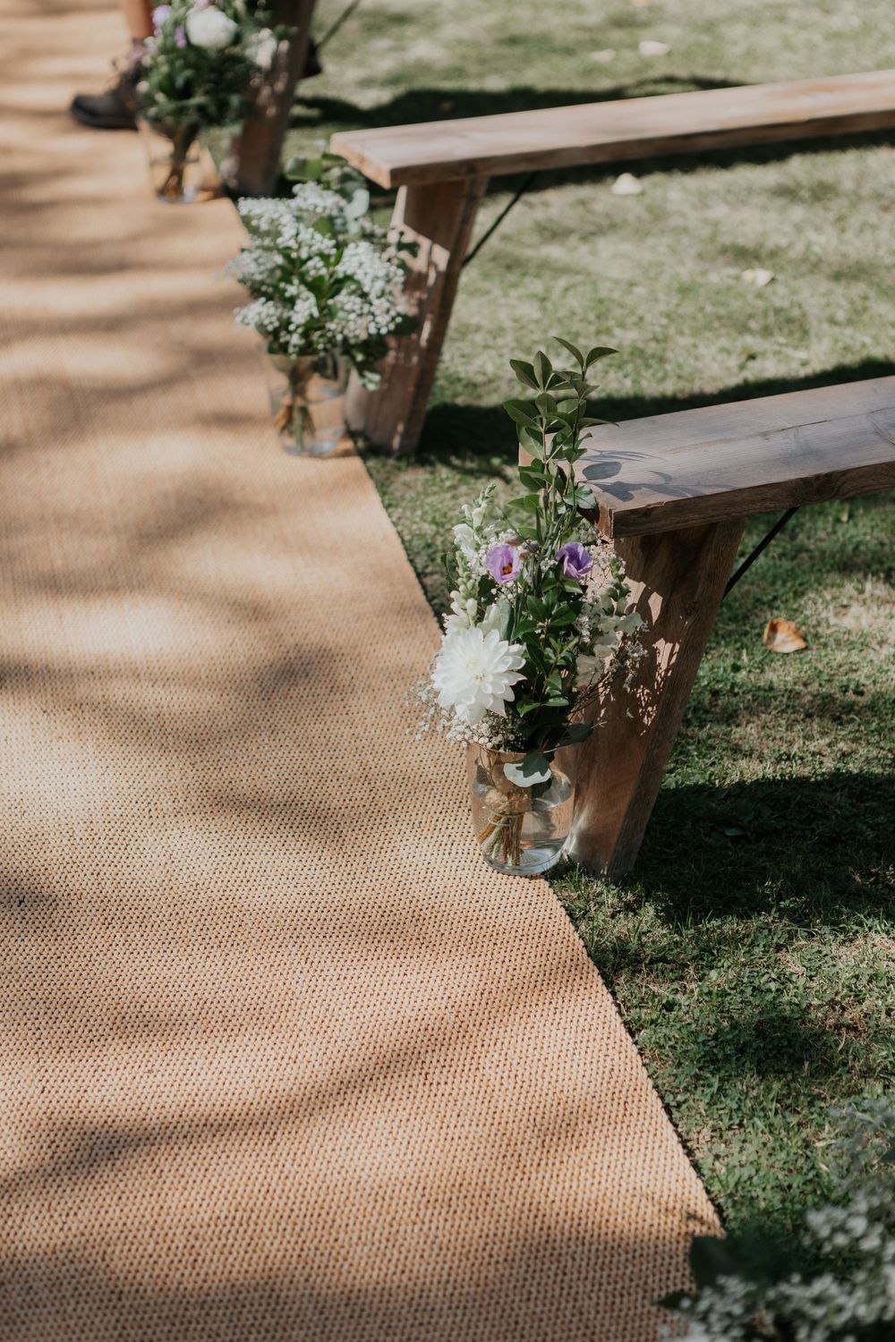Rustic outdoor wedding ceremony aisle with burlap runner and floral jar decorations on benches.