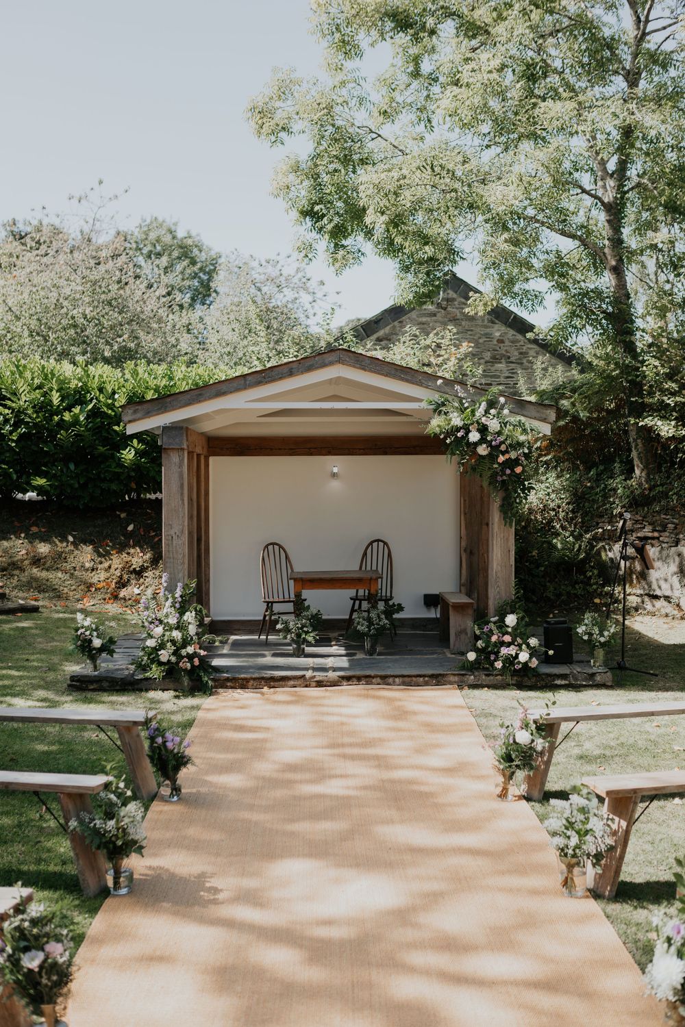 Outdoor wedding ceremony setup with floral arch, wooden benches, and burlap aisle runner.