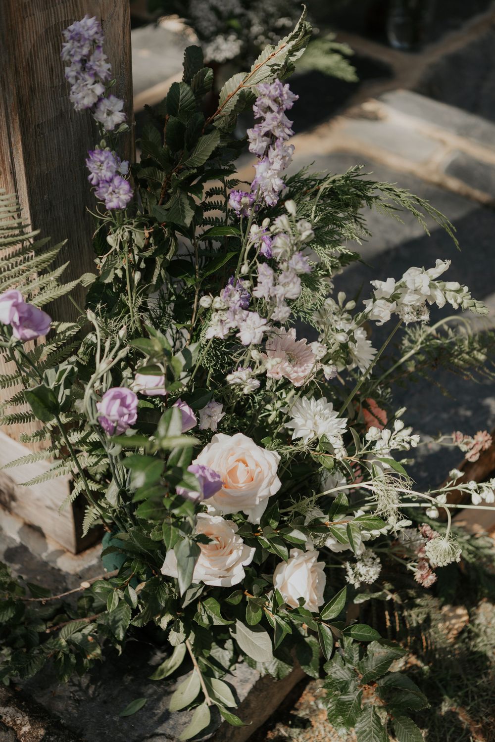 Wild garden-style wedding flowers with roses, purple delphiniums, and ferns at ceremony entrance.