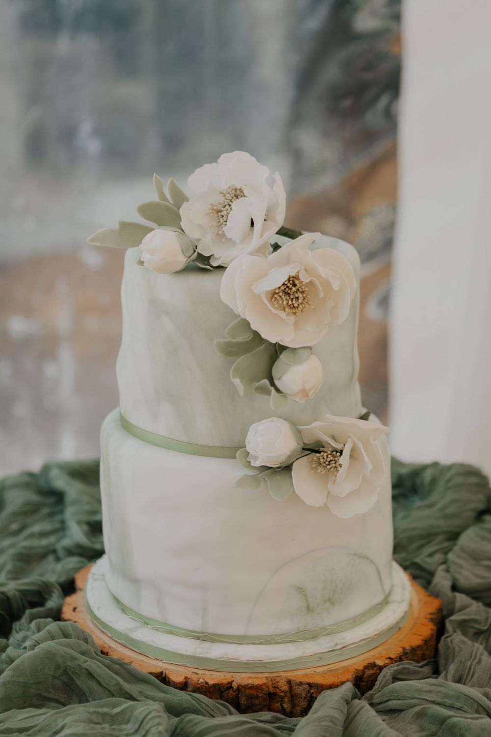 Elegant two-tier white and sage green marble wedding cake adorned with white peony flowers and green leaves.
