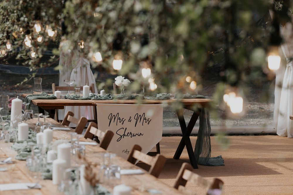 Rustic outdoor wedding reception table with Mr & Mrs sign, candles, and hanging string lights among trees.