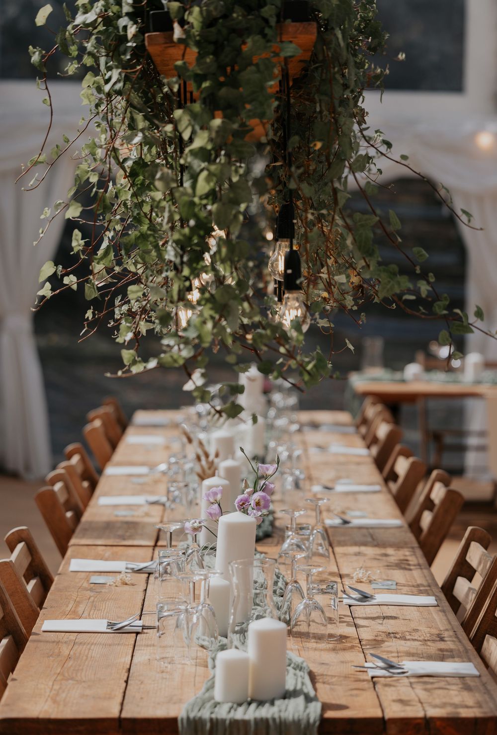 Long rustic wooden wedding reception table with white candles, greenery runner, and lush hanging botanical installation.