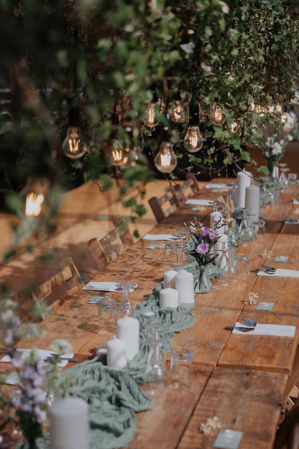 Romantic outdoor wedding table setting with Edison bulb lights, greenery overhead, white candles, and purple flowers.