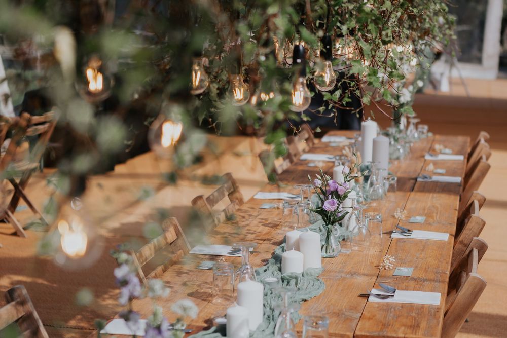 Rustic wooden wedding banquet table with sage green runner, white candles, floral centerpieces, and Edison bulb lights.