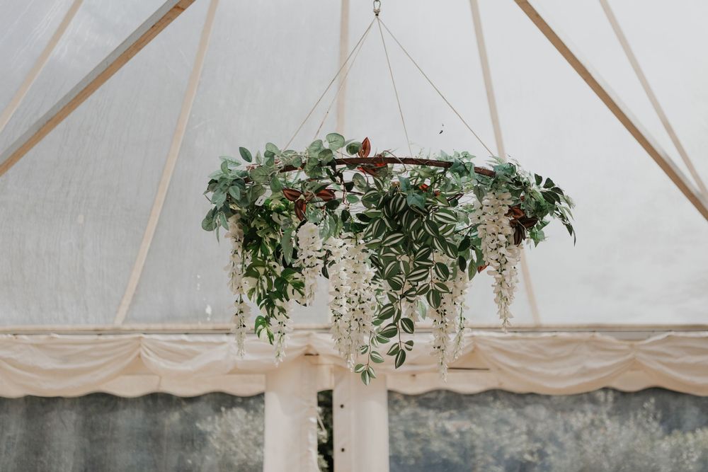 Elegant hanging floral chandelier with white wisteria and greenery inside a marquee tent.