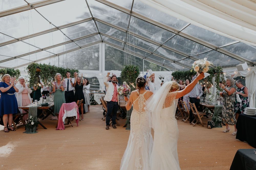 Two brides enter a clear marquee reception tent as guests applaud and celebrate their wedding arrival.