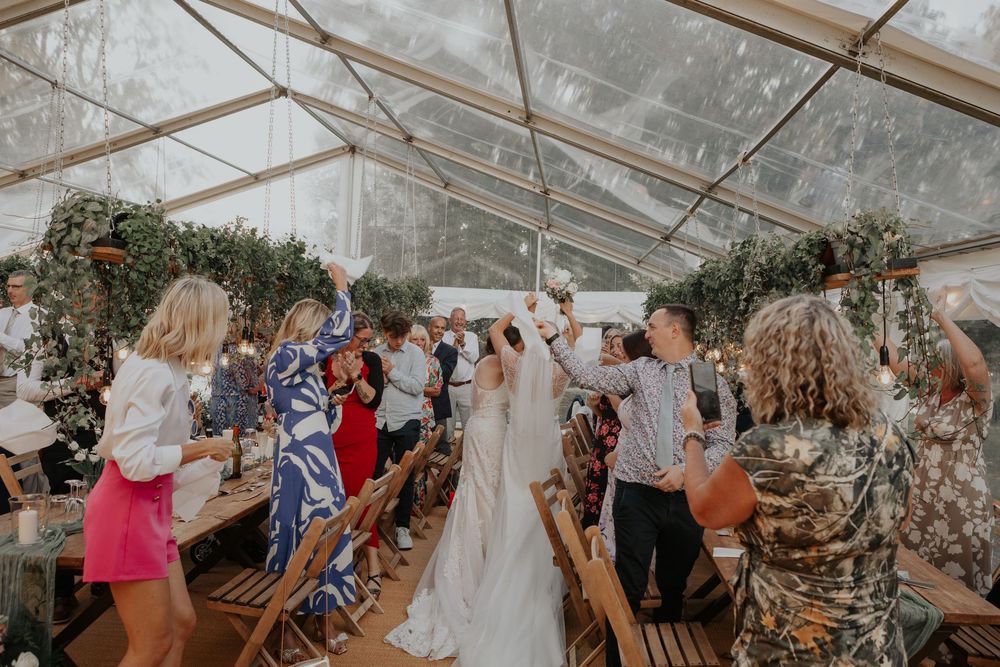 Two brides walk through a cheering crowd inside a clear-roofed marquee during their wedding reception.