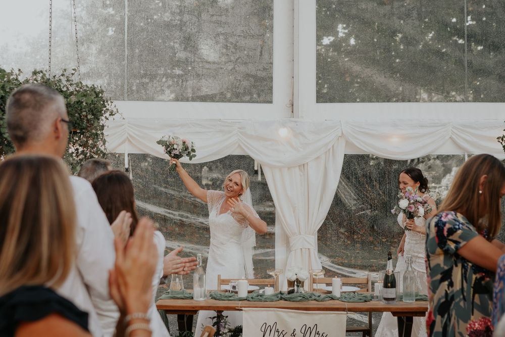 A bride raises her bouquet triumphantly at the wedding reception table decorated with Mrs and Mrs signage.