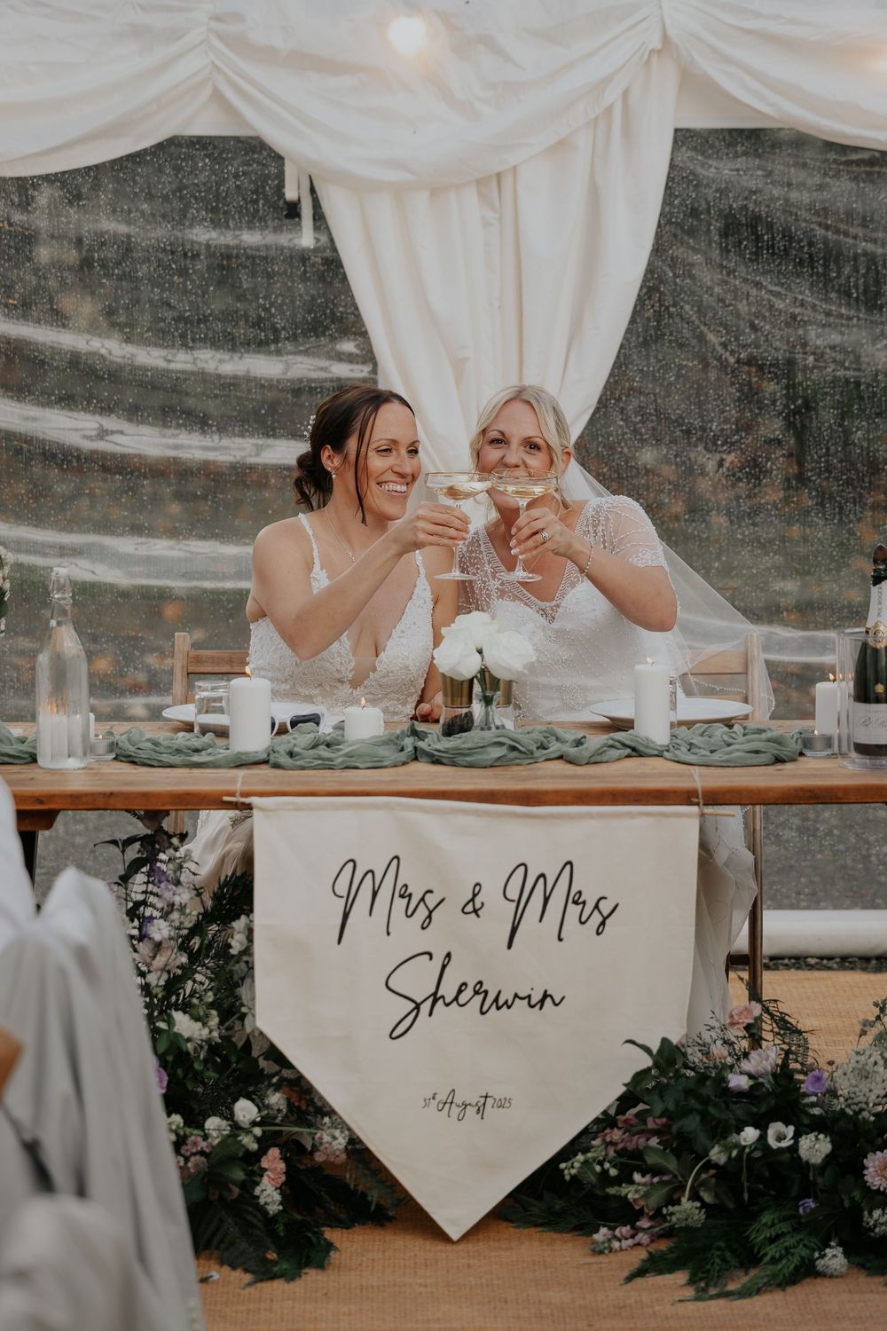 Two brides toast champagne at wedding reception table with Mrs & Mrs Sherwin banner and floral decorations.