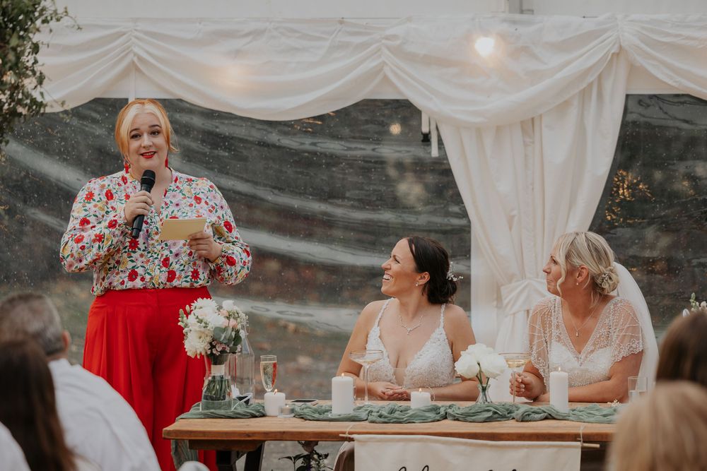 Wedding guest in floral top and red skirt gives speech while two brides laugh at reception table.