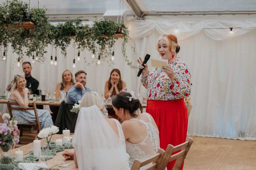 Red-skirted wedding speaker addresses smiling guests in elegant marquee with hanging greenery decor.
