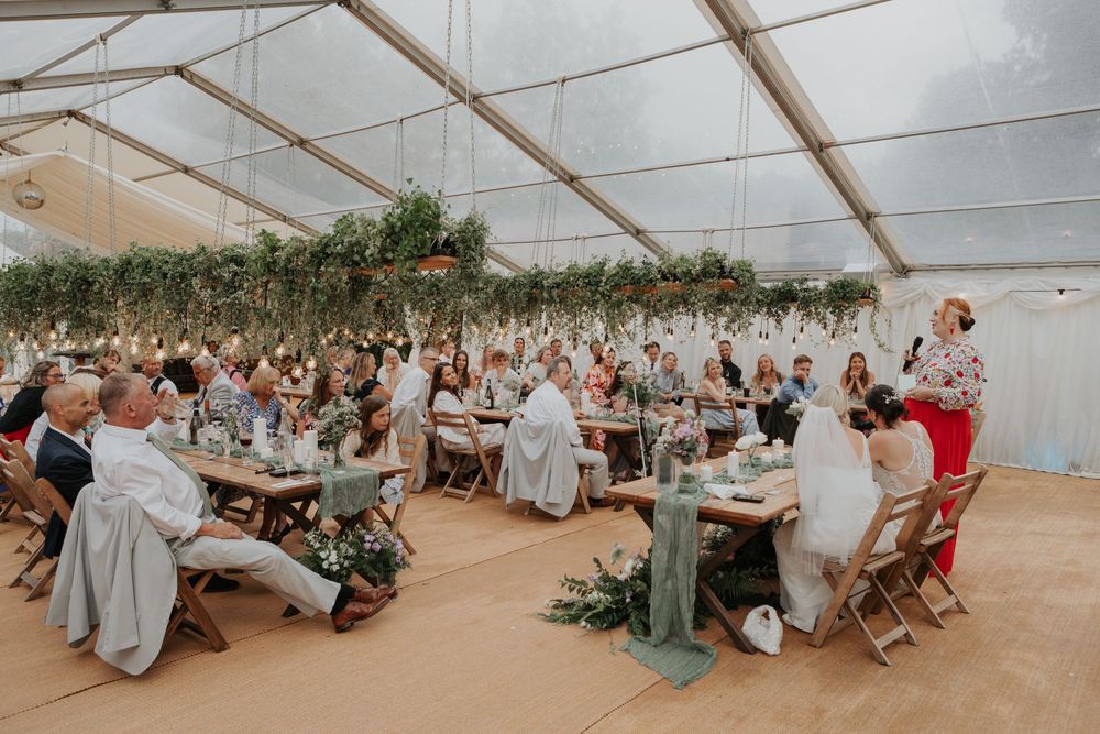 Wide shot of wedding reception in clear marquee with long wooden tables and hanging Edison bulb lights.
