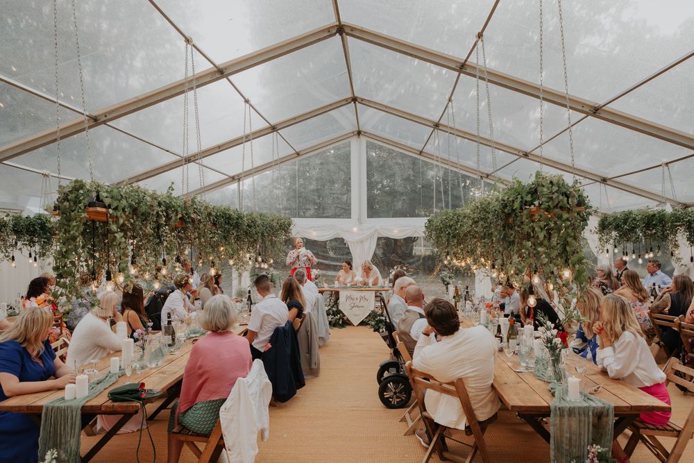 Aerial view of elegant wedding reception in glass marquee with greenery ceiling and warm Edison lighting.