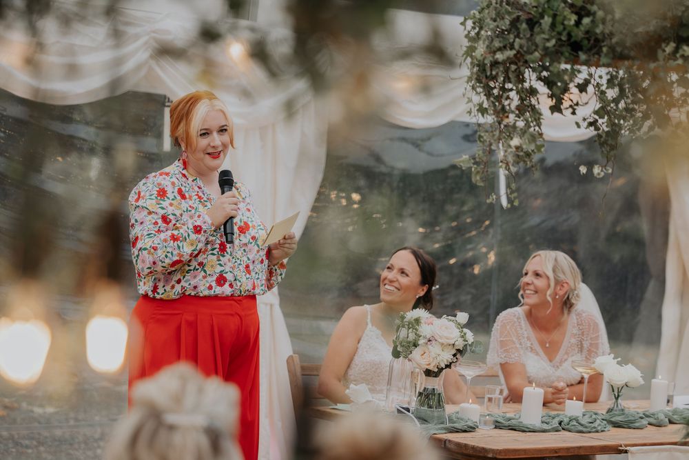 Wedding speech moment with brides laughing at head table under fairy lights in outdoor marquee.