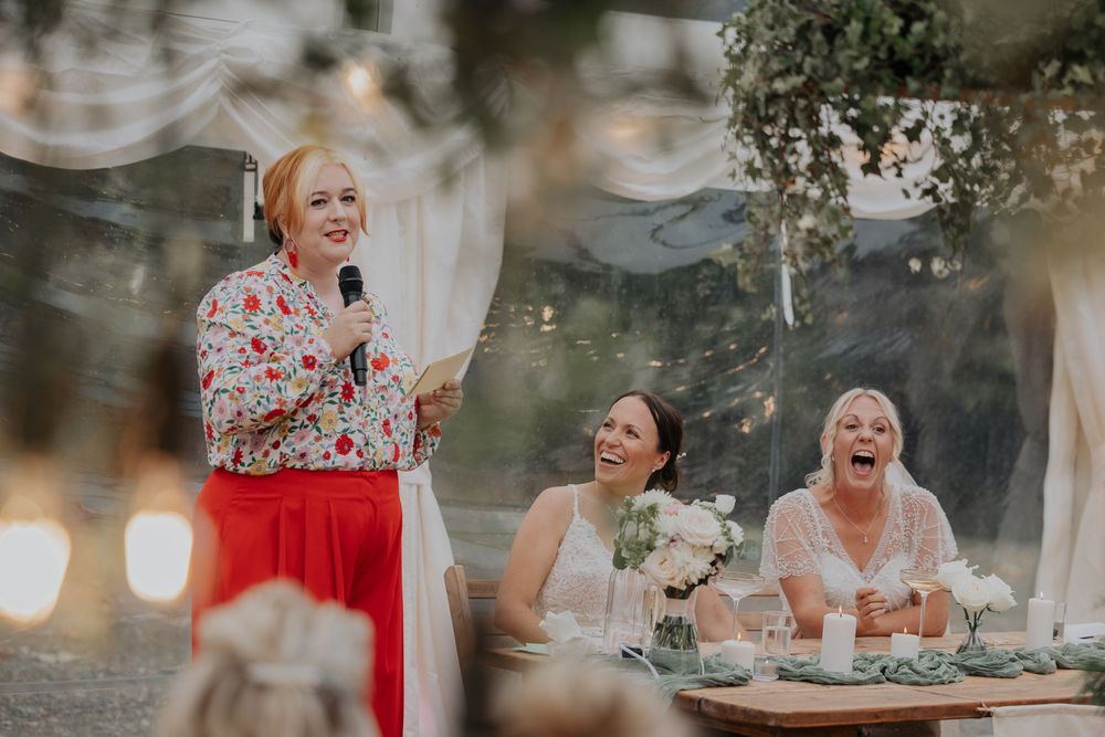 Brides laugh joyfully during wedding speech in decorated marquee with string lights and white draping.