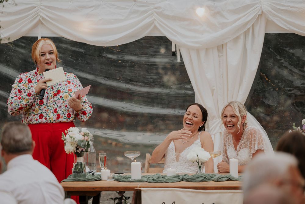 Wedding speaker reads from notes while brides smile at floral-decorated reception table in marquee.